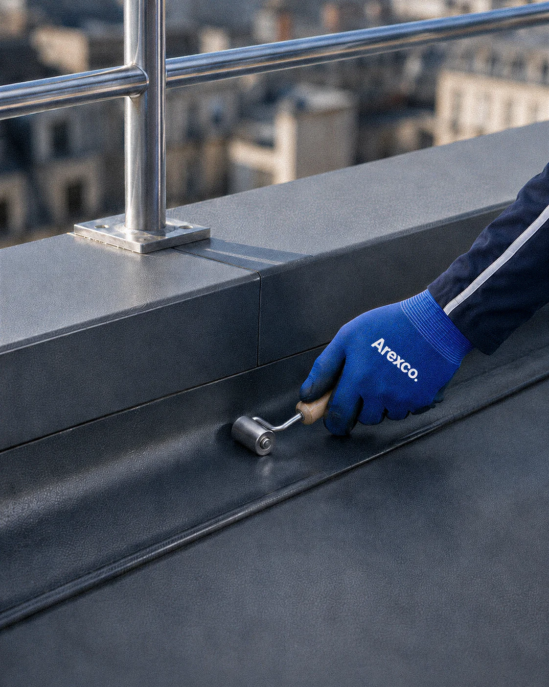 Main gantée appliquant une membrane d'étanchéité au rouleau sur une toiture-terrasse moderne en réhabilitation.